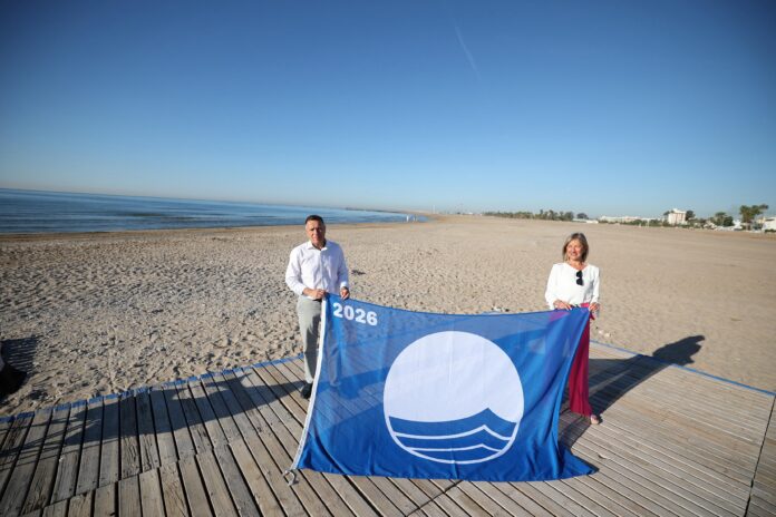 Man and woman on a sunny beach holding a large blue banner with a white waves logo and the year 2026 visible on the banner.