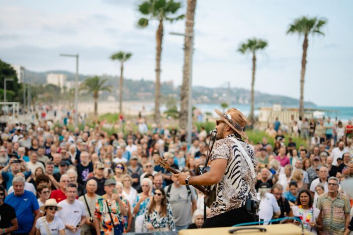 Musician with hat and patterned shirt playing guitar at an outdoor beach concert as a large crowd watches nearby.
