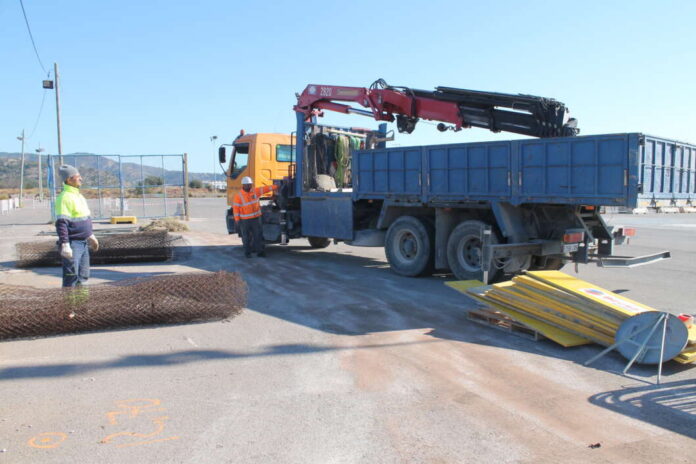 Truck with a crane-mounted lifting arm on a construction site; two workers in high‑visibility vests nearby.