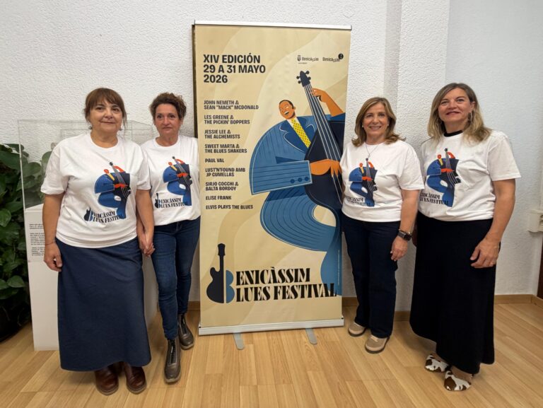 Four women in white festival T-shirts pose beside a tall Benicàssim Blues Festival poster indoors.