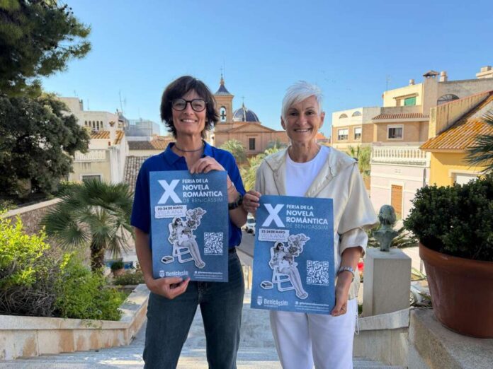 Two smiling women stand on a terrace holding blue posters promoting the Benicàssim Romance Novel Fair.