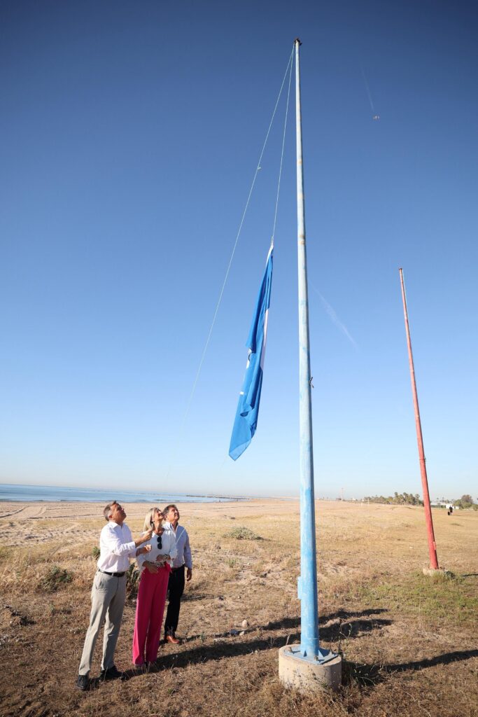Three adults on a beach gaze upward as a blue flag is raised on a tall pole against a clear blue sky.