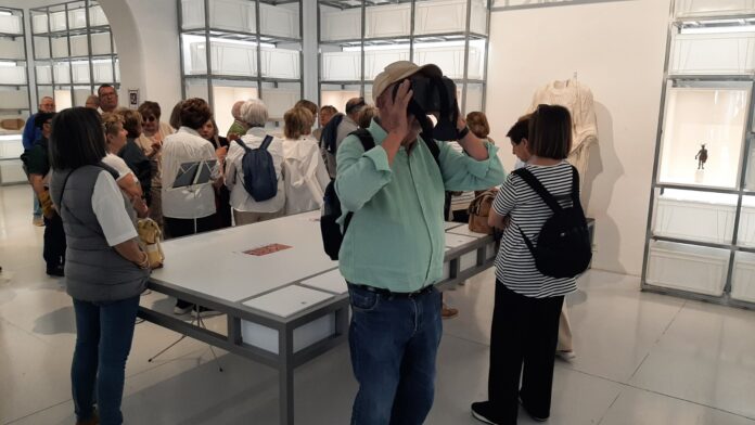 Man wearing a VR headset, surrounded by museum visitors in a bright gallery with display cases and a statue exhibit behind him.