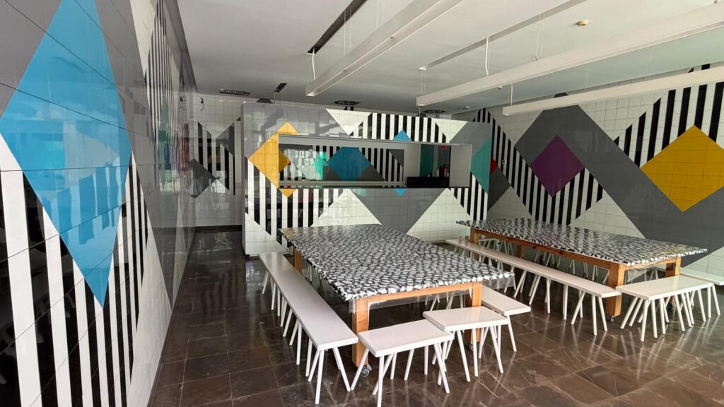 A bright cafeteria with geometric wall art—black-and-white stripes and large colorful diamonds—around long tables with patterned black-and-white tablecloths and white benches.