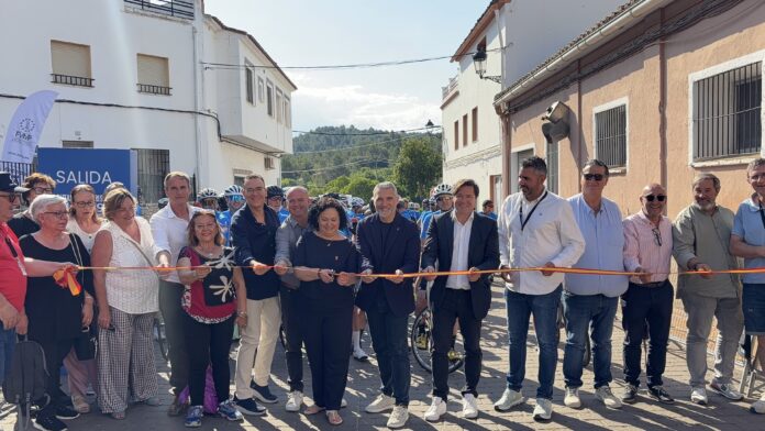 Group of adults and spectators standing along a street, holding an orange ribbon for a ribbon-cutting ceremony outside white and peach buildings.