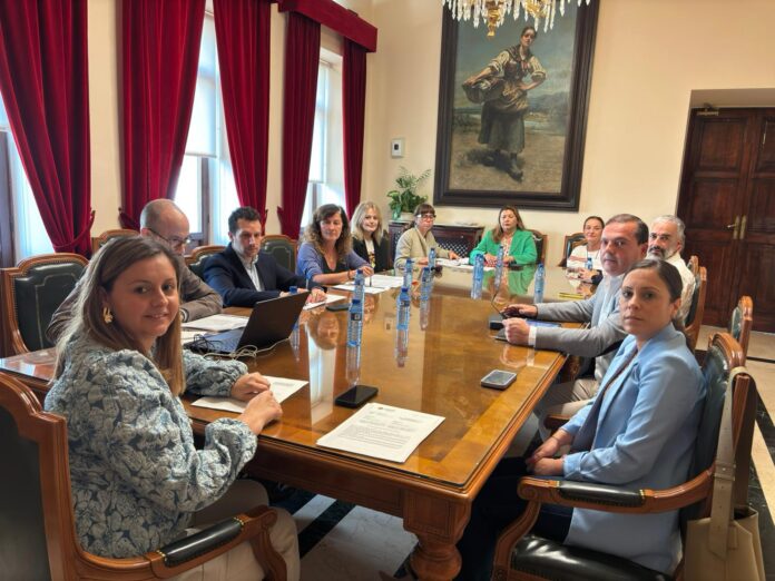 Ten professionals sit around a polished wooden conference table in a room with red drapes and a large framed painting, engaged in a meeting.