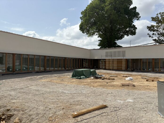Construction site in front of a modern, single-story white building with large glass windows and wooden frames; a tall tree rises behind.