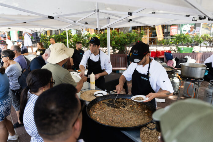 Cooks prepare a large paella under a white canopy while a crowd watches at an outdoor market show.