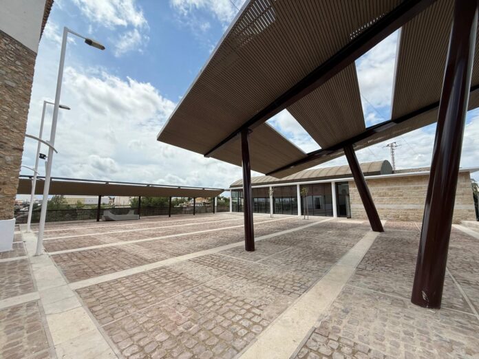 Open-air plaza with large slatted wooden canopies supported by dark brown columns over a cobblestone surface, cloudy sky visible above.