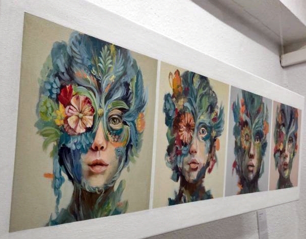 Series of five portrait panels mounted on a white wall, each showing a woman's face partially obscured by a bright, floral-patterned mask.