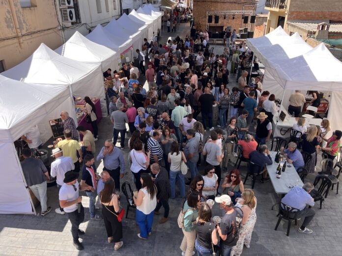 Crowded outdoor market with white vendor tents lining a narrow street and people browsing stalls under sunny skies.
