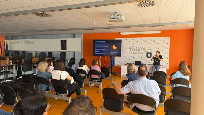 Presenter speaks at a podium in a bright conference room, with a projected slide and an attentive audience seated in rows behind him.