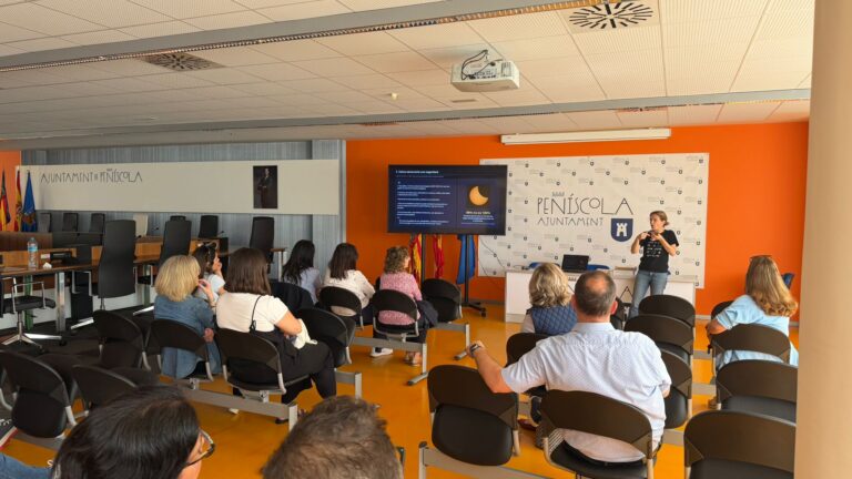 Presenter speaks at a podium in a bright conference room, with a projected slide and an attentive audience seated in rows behind him.