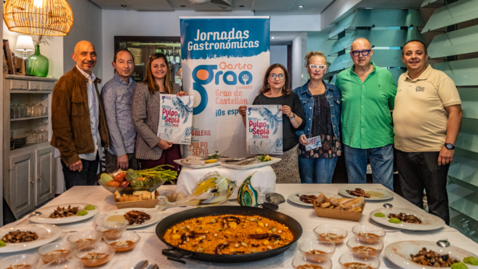 Eight people stand behind a long table of dishes at a gastronomic event, with a blue 'Jornadas Gastronomicas' banner in the center and various foods displayed front.