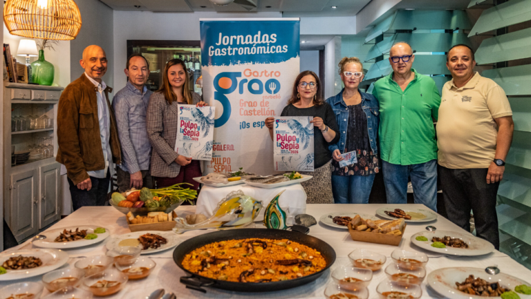 Eight people stand behind a long table of dishes at a gastronomic event, with a blue 'Jornadas Gastronomicas' banner in the center and various foods displayed front.
