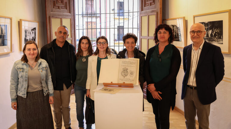Group of eight adults posing in an art gallery near a display pedestal with framed photos on the walls behind them