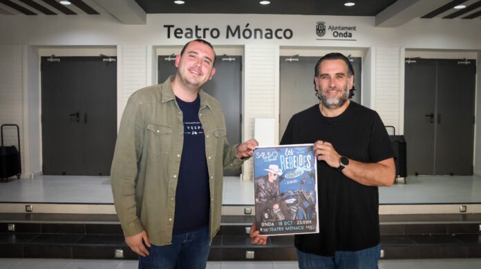 Two men stand in a theater lobby, smiling, while holding a colorful poster for a show in front of a Teatro Mónaco sign.