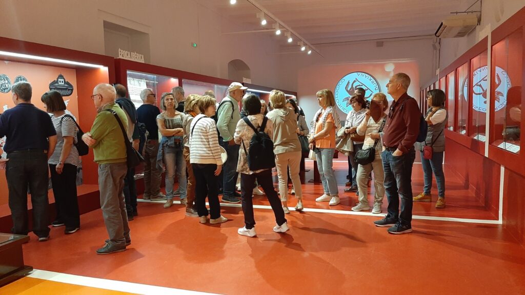 Group of visitors in a red-walled museum gallery, looking at displays along glass cases and wall panels.