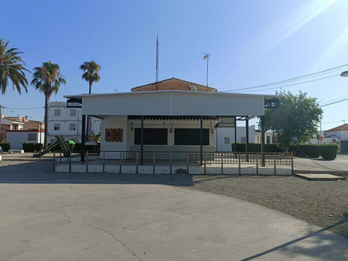 Two-story white building with a gated front porch, palm trees on the left, and tall antenna masts on the roof against a clear blue sky.