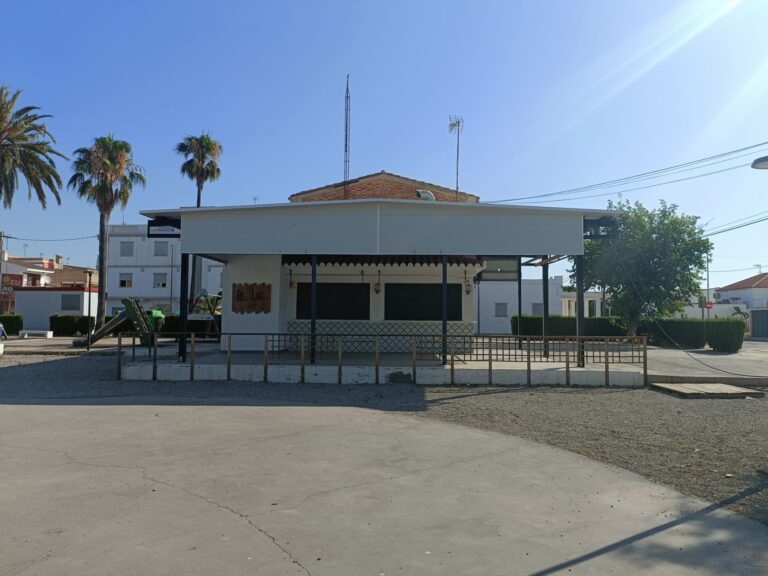 Two-story white building with a gated front porch, palm trees on the left, and tall antenna masts on the roof against a clear blue sky.