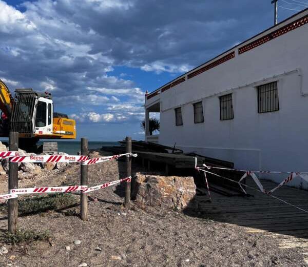 Coastal construction site with a yellow excavator beside a white building, red-and-white safety tape, and a wooden ramp over rocky ground.