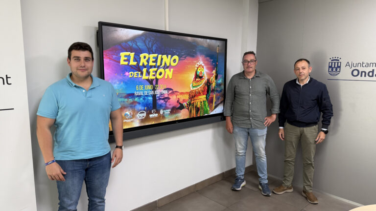 Three men stand beside a large colorful screen advertising 'El Reino del Leon' with event details at a municipal office backdrop (Ajuntament Onda).