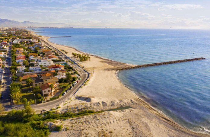 Aerial view of a coastal town with a sandy beach, calm blue sea, and a long breakwater jetty extending into the water.