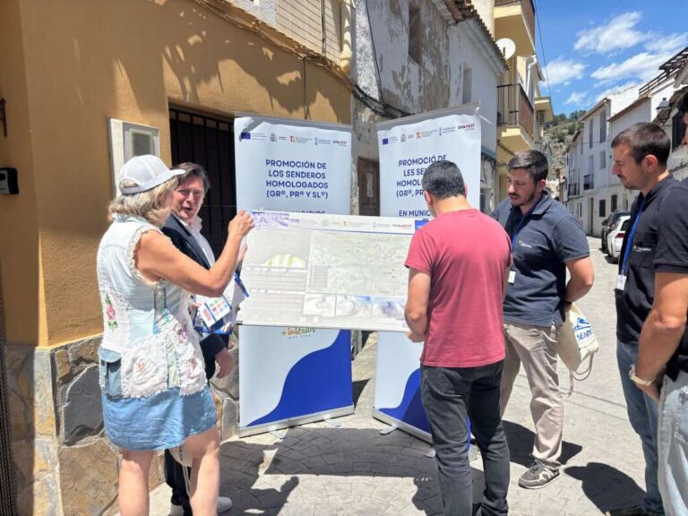 Group of five adults standing on a sunlit street, examining a large map display with banners about promoted trails at an outdoor info stand.