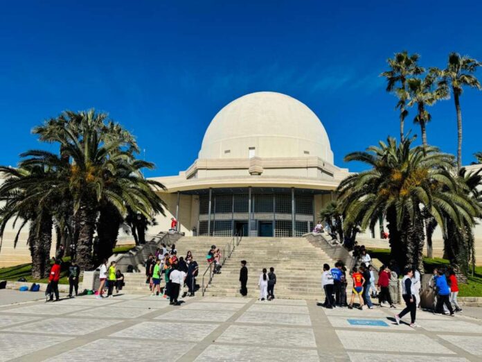 Group of people gathered outside a large domed building with palm trees and a bright blue sky in the background.