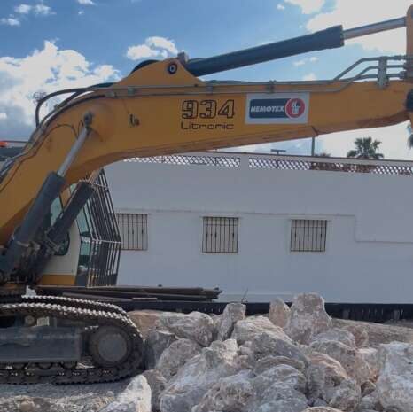Close-up of a yellow excavator arm with the number 934 and 'Liebherr' branding, resting on a pile of rocks at a construction site with a white building in the background.