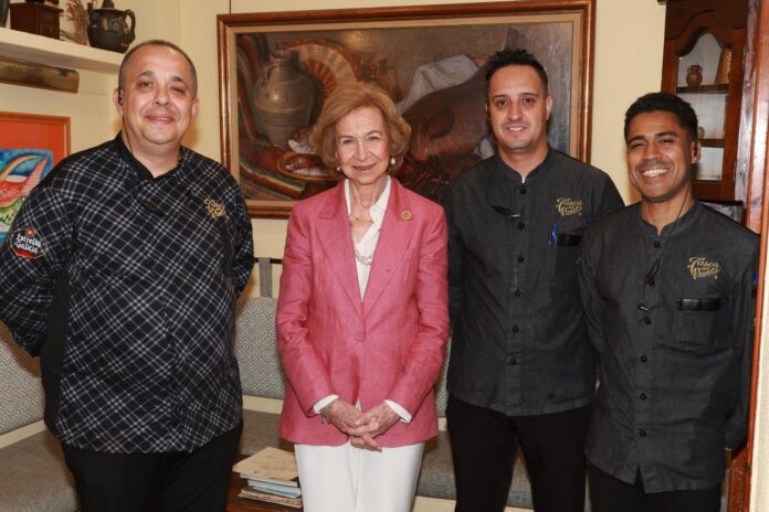 Four people pose for a photo in a warmly lit room: woman in a pink blazer center, flanked by two servers in dark uniforms and a chef in a plaid jacket on the left, with a framed painting behind them.