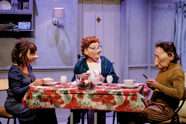 Three puppet characters sit around a dining table with a red floral tablecloth, eating bowls in a theater stage kitchen set.