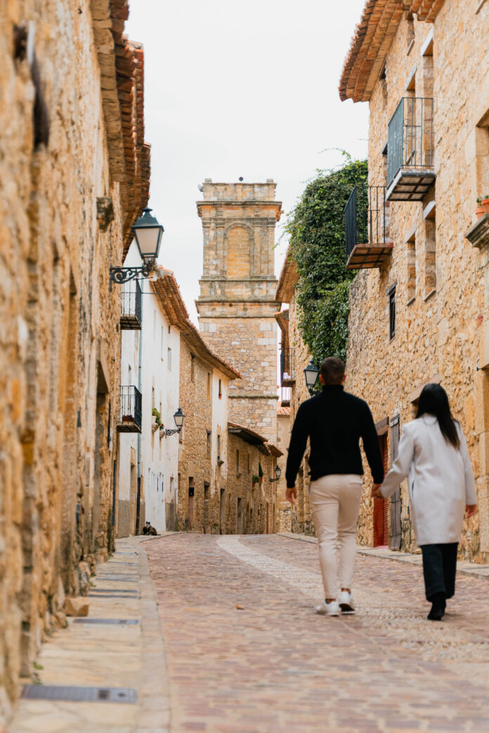 Two people walk away down a narrow cobblestone street flanked by stone buildings with balconies and a distant historic tower.