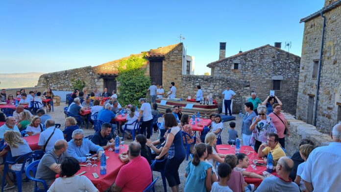 Outdoor communal meal in a stone courtyard with red-tablecloth tables and many guests dining together on plastic chairs.