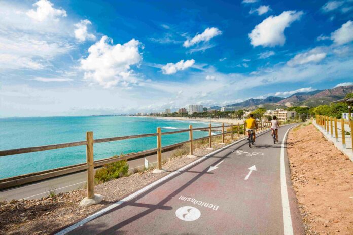 Two cyclists ride along a coastal bike path beside the turquoise sea, with a wooden railing and distant city buildings under a blue sky.