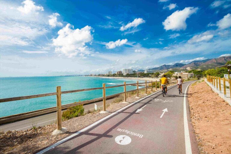 Two cyclists ride along a coastal bike path beside the turquoise sea, with a wooden railing and distant city buildings under a blue sky.