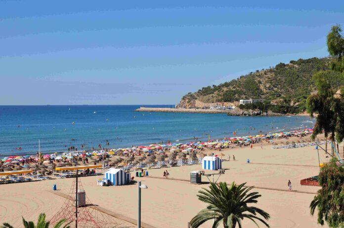 Busy sandy beach with colorful umbrellas and sunbathers, calm blue sea and a rocky hillside to the right under a clear sky.