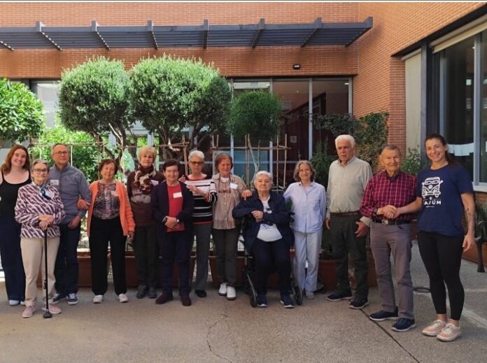 Group of seniors posing for a group photo outside a brick building with glass doors; some stand, one uses a wheelchair.