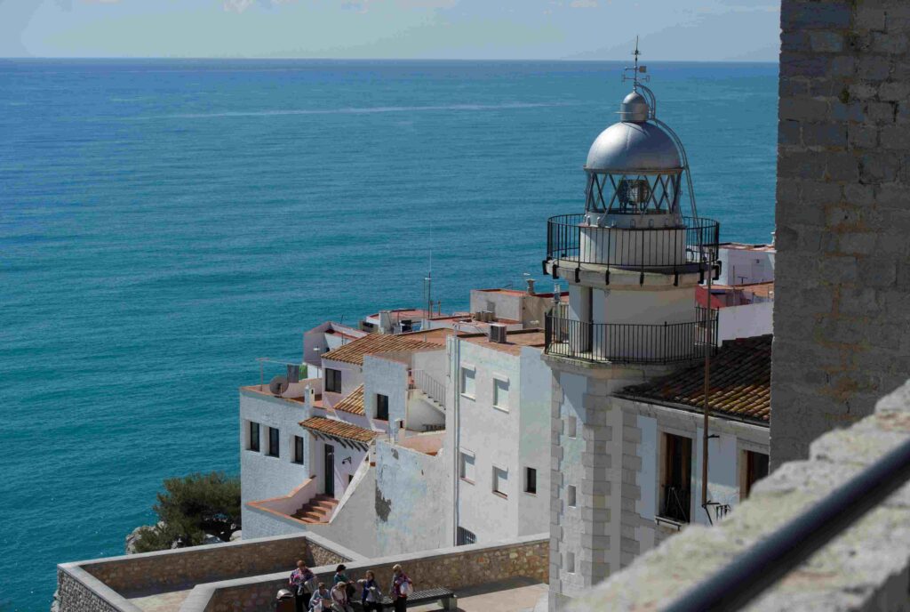 Coastal town with white buildings and a domed lighthouse overlooking a blue sea, with people gathered on a stone terrace below.