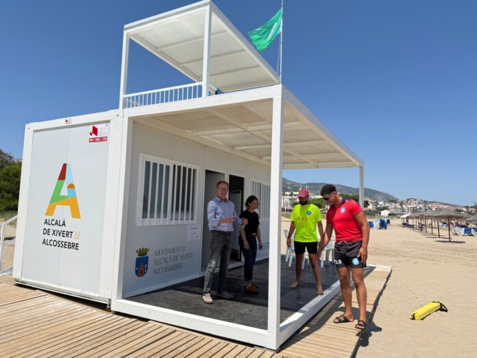 Beach information booth on a wooden boardwalk at the seaside, with three people standing in front and a green flag flying above the structure.
