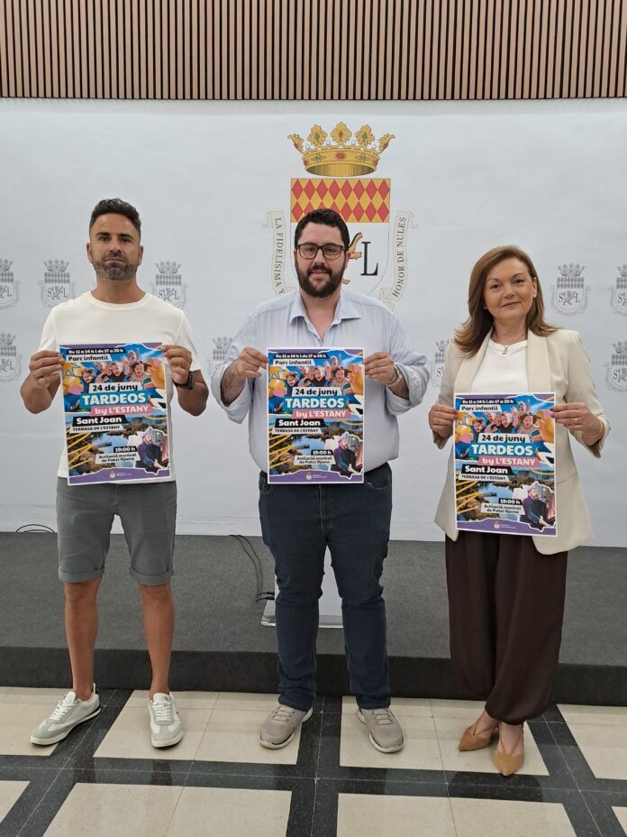 Three adults stand indoors in front of a crest backdrop, each holding a colorful poster for a June 24 event.