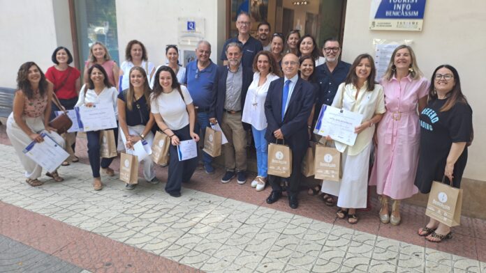 Group of adults posing outside a building, holding certificates and Benicàssim gift bags, smiling for a photo during a ceremony in daylight.