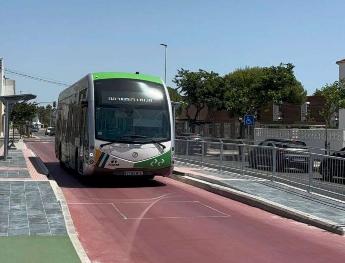 Front view of a modern green-topped tram traveling on a red transit lane with trees and parked cars in the background.