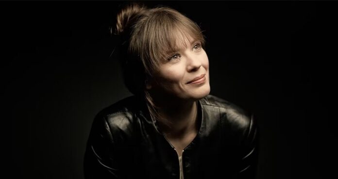 Portrait of a woman with light hair in a black leather jacket, looking upward with a soft smile against a dark background.