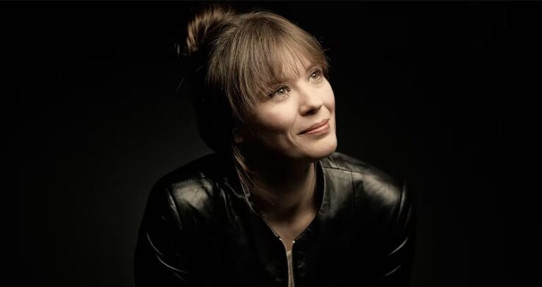 Portrait of a woman with light hair in a black leather jacket, looking upward with a soft smile against a dark background.