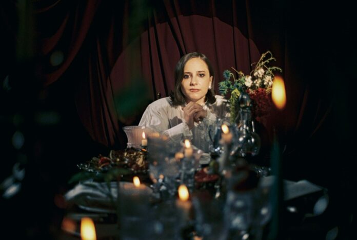 Woman with dark hair sits at a candlelit dining table, chin resting on hands, looking at the camera.