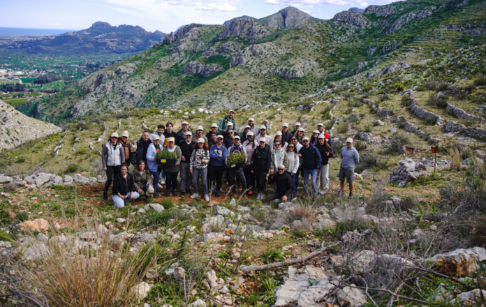 Large group of people wearing hats posing on a rocky hillside with terraced slopes and mountains in the distance.