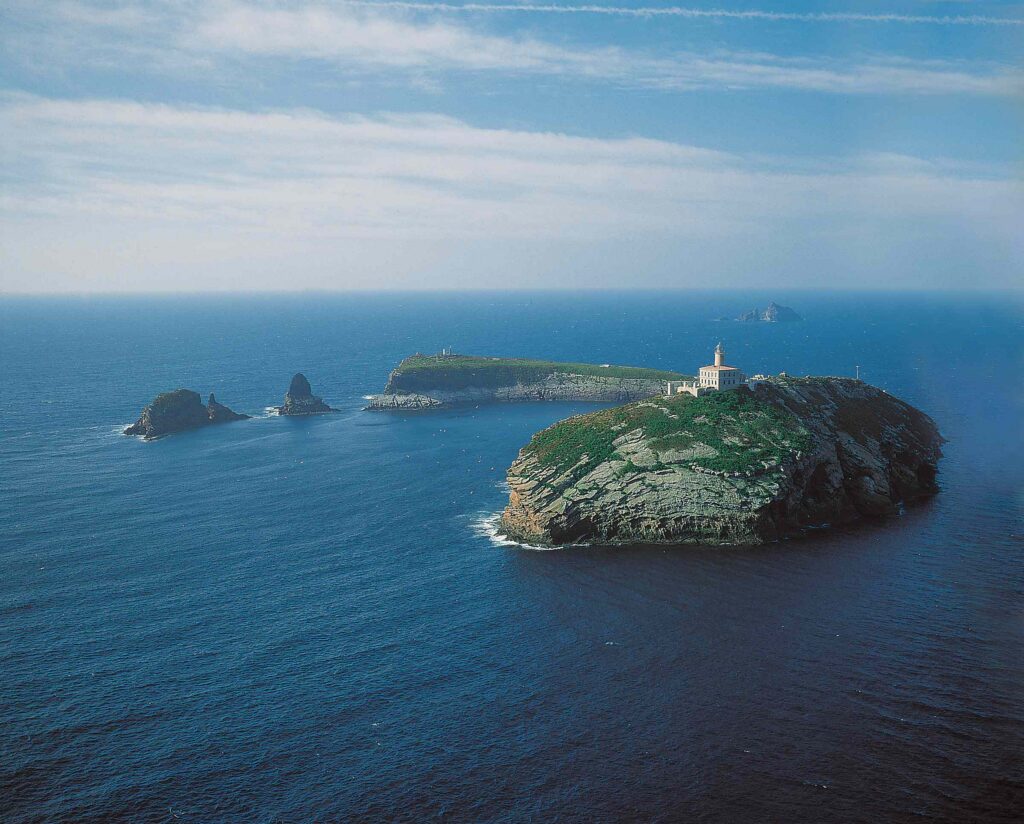Lighthouse perched on a rocky coastal headland overlooking the blue sea and a distant island.