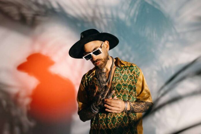 Man wearing a wide-brimmed black hat, white sunglasses, and a green-gold patterned shirt, with visible tattoos and jewelry, posing against a blurred backdrop.
