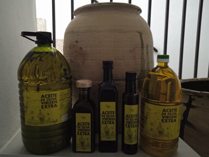 Shelf with multiple olive oil bottles, labels read 'ACEITE DE OLIVA VIRGEN EXTRA'; large green bottle left, clear glass bottle right, dark bottles in between.